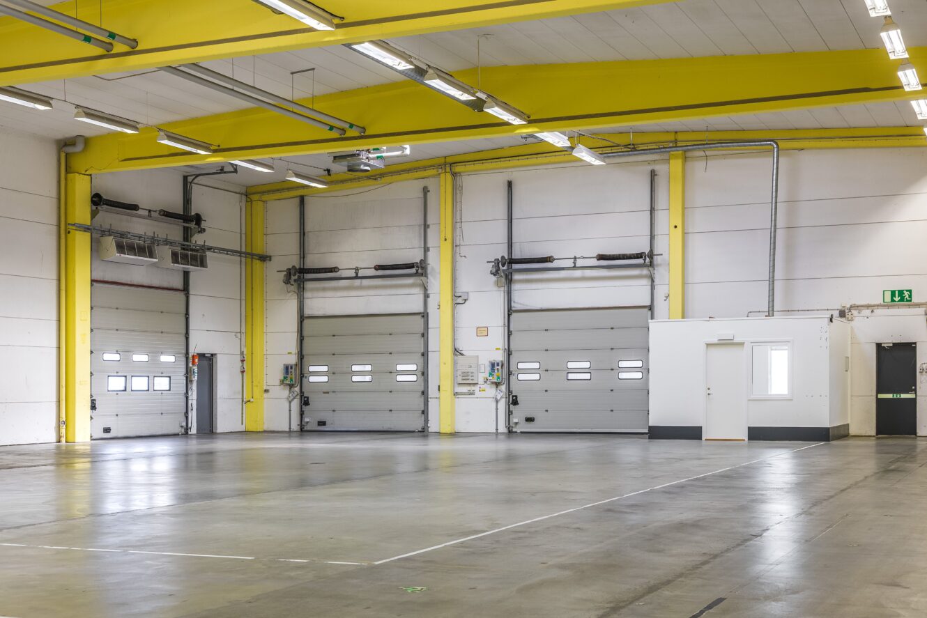 Tom lagerlokal med tre stora grå garageportar, högt i tak med gula balkar, takbelysning och ett litet vitt kontorsutrymme till höger.
