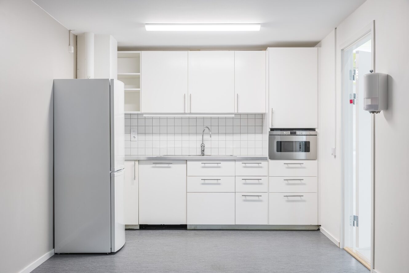 Minimalistiskt vitt kök med skåp, kylskåp, diskho, spishäll och inbyggd mikrovågsugn mot ett kaklat stänkskydd under stark takbelysning.