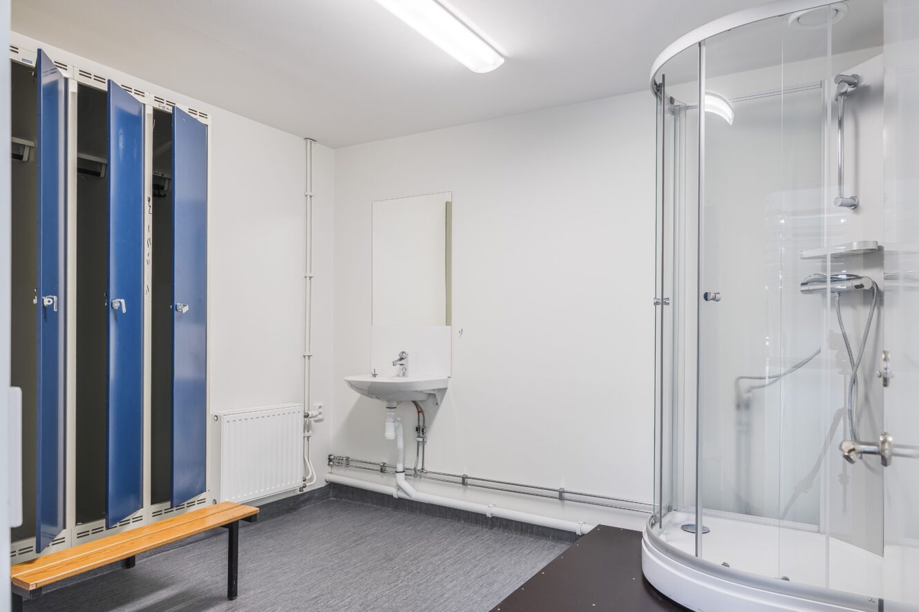 Omklädningsrum med blå skåp, en träbänk, ett vitt handfat med spegel och en dusch med glasväggar på ett grått golv.