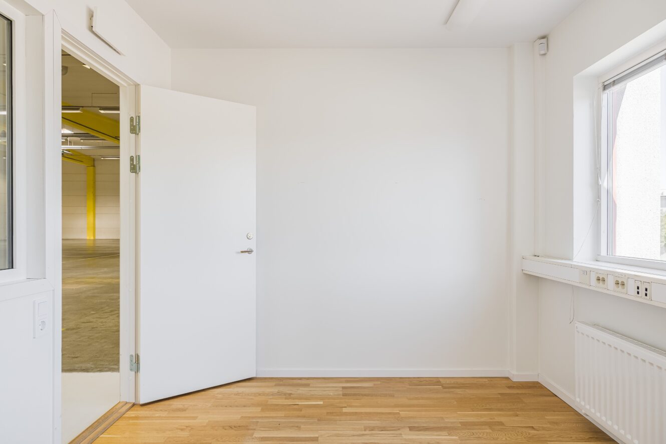 Tomt vitt rum med ljust trägolv, öppen dörr som leder till ett industriområde, stort fönster och eluttag på väggen.