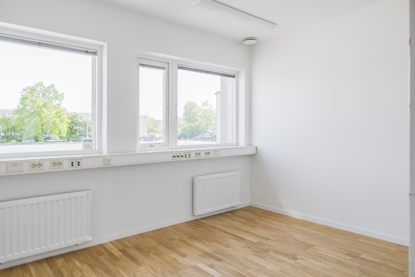 Tomt, omöblerat rum med vita väggar, stora fönster som släpper in naturligt ljus, trägolv och radiatorer under fönstren.