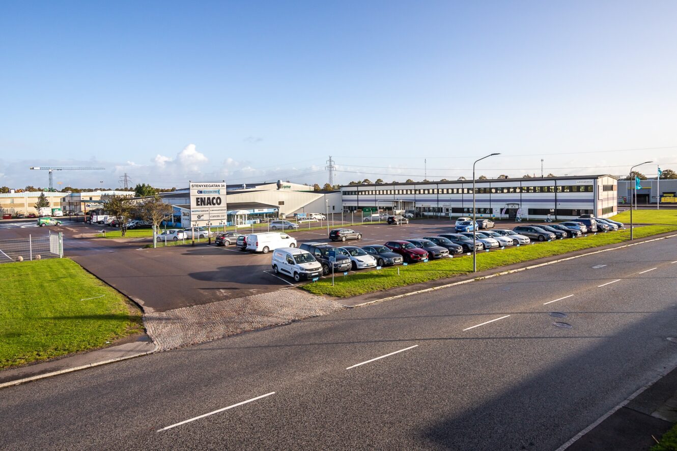 En kommersiell bilhall med många parkerade bilar, en byggnad märkt ENACO och en klar himmel i bakgrunden.