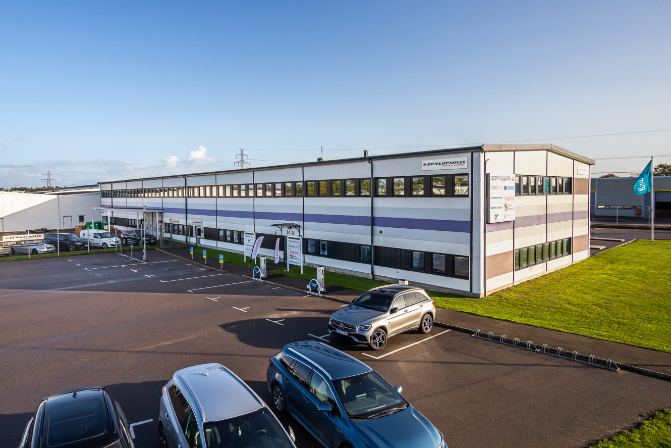 En modern kontorsbyggnad med stora fönster och en parkeringsplats med flera bilar en klar dag.