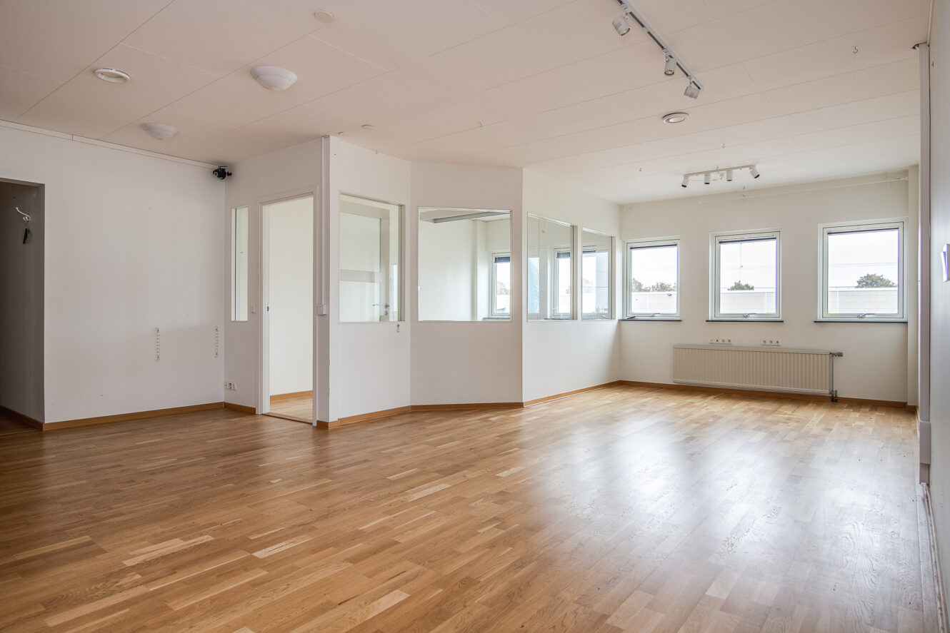 Rymligt tomt rum med ljust trägolv, vita väggar, flera fönster, taklampor och ett litet slutet glasparti på ena sidan.