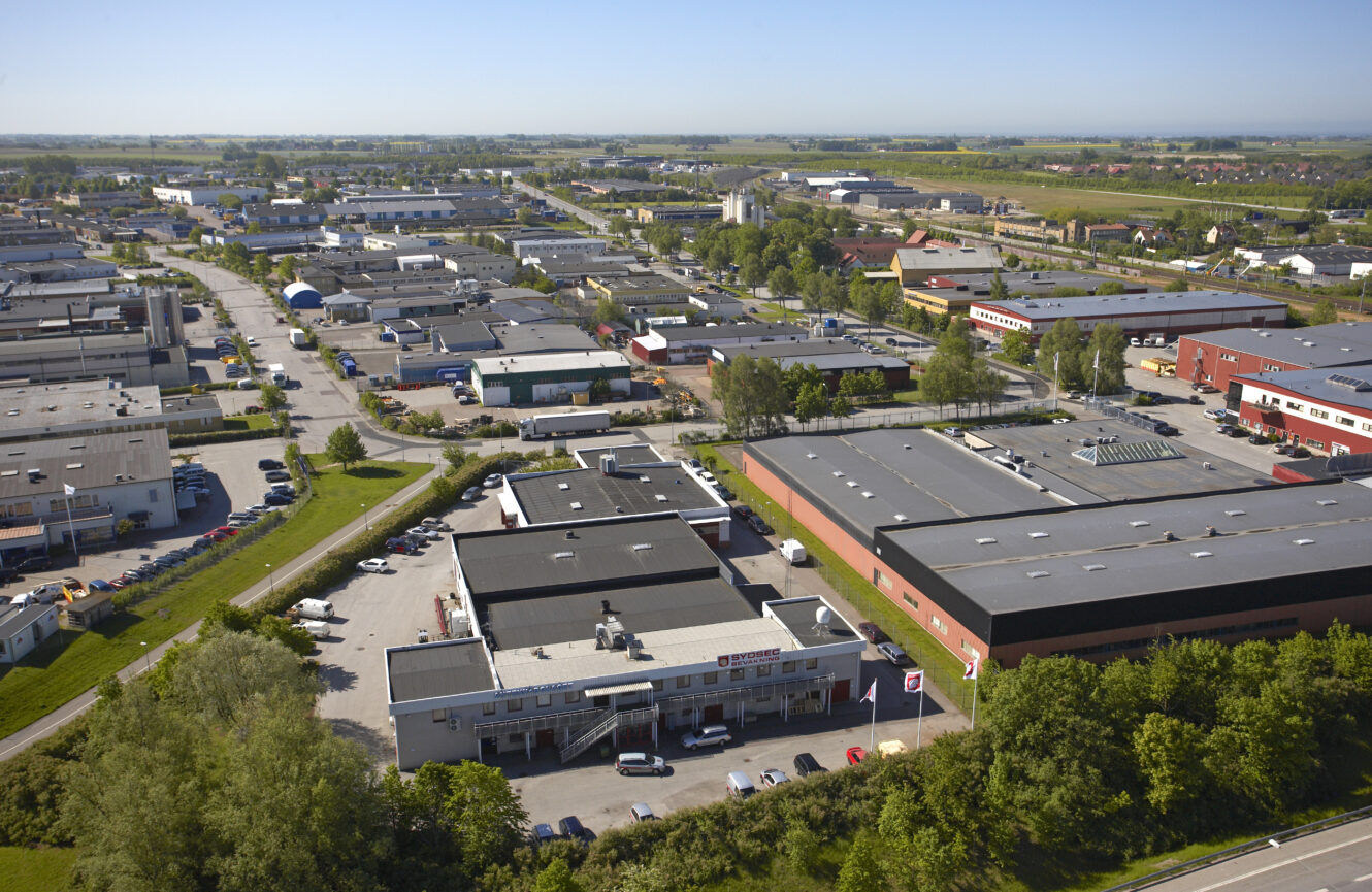 Flygfoto över en industripark med flera stora lagerbyggnader, vägar och grönområden under en klarblå himmel.