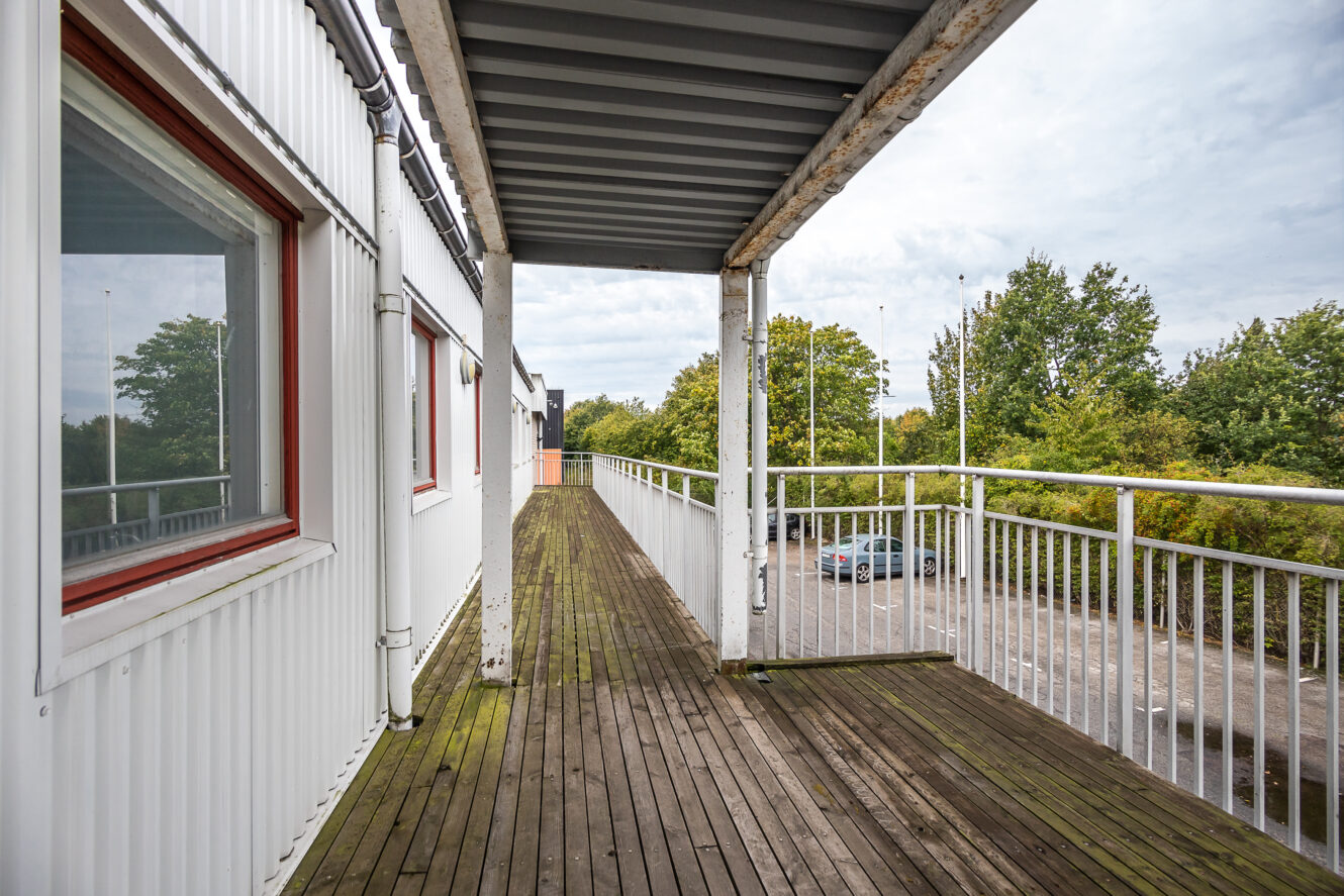 En lång, upphöjd gångväg i trä med metalltak och räcke, som är kopplad till en vit byggnad, har utsikt över en parkeringsplats och träd.