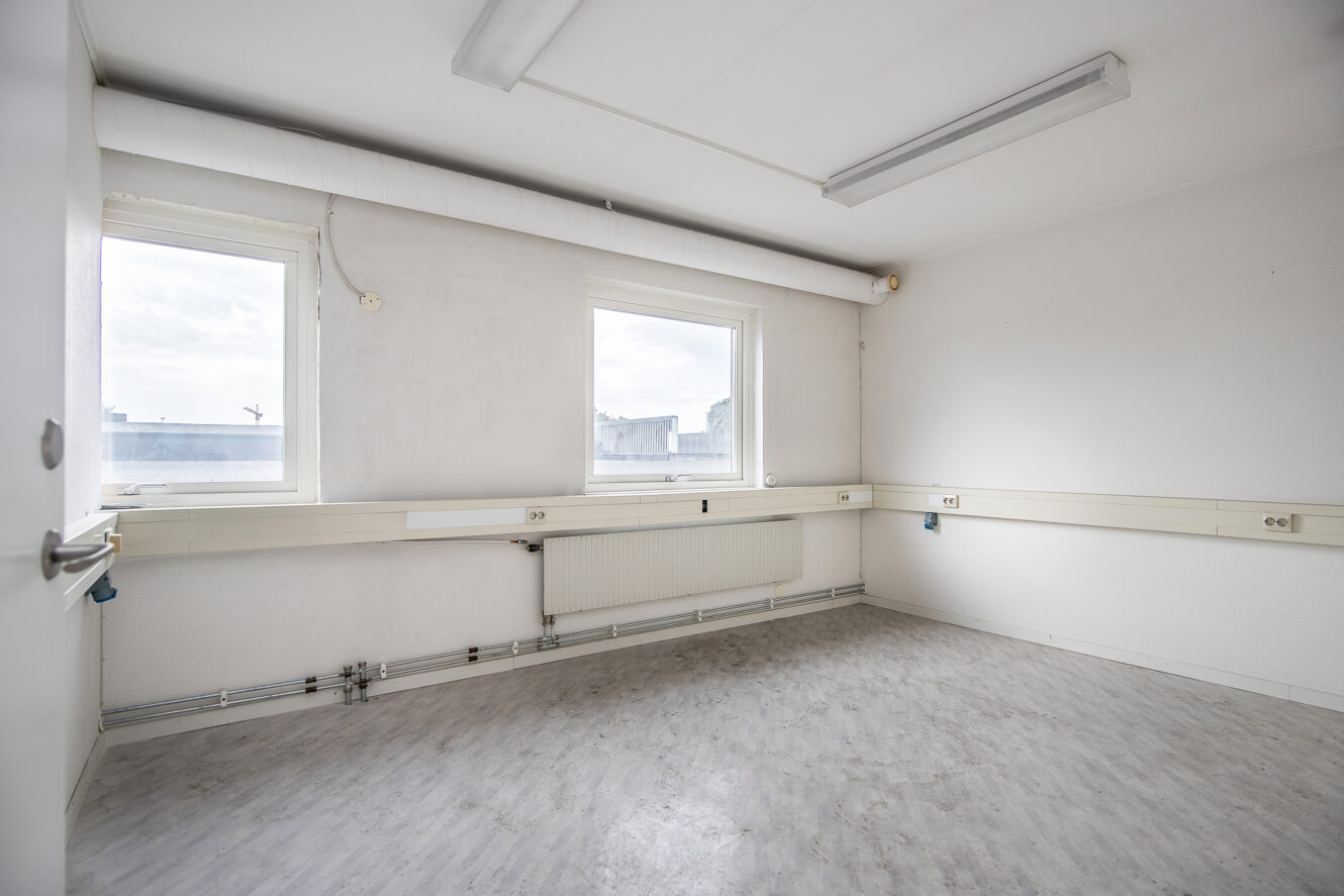 Tomt, omöblerat rum med vita väggar, två fönster, lysrör i taket och ljusgrått golv. Eluttag längs väggarna.