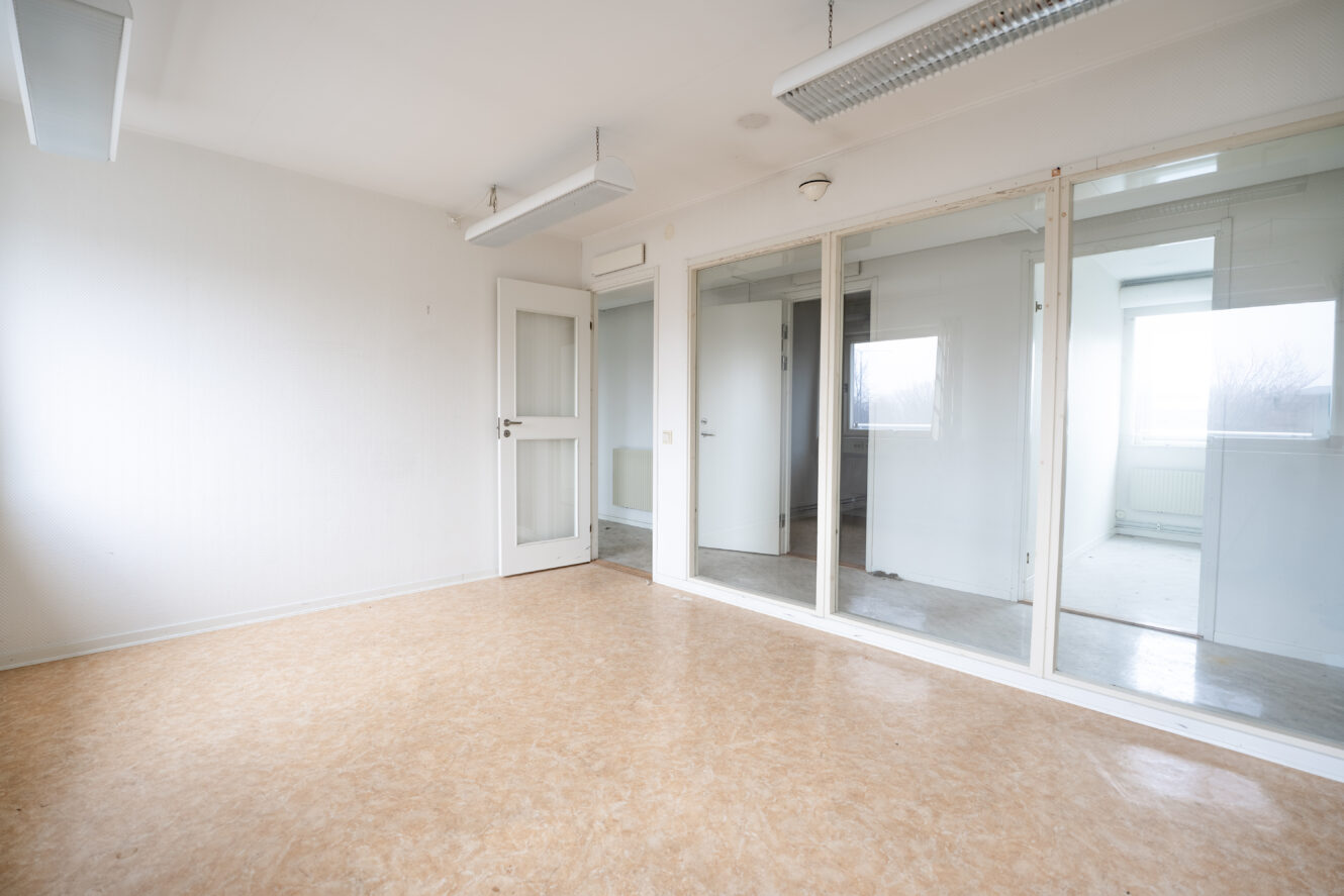 Tomt kontorsrum med beige linoleumgolv, vita väggar, lysrör i taket, en stängd dörr och stora innerfönster med utsikt över ett annat rum.