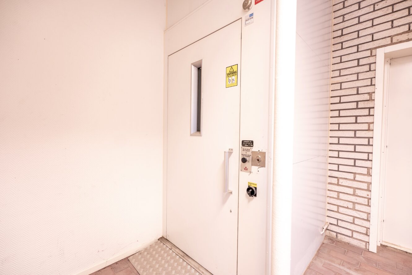 En vit hissdörr med ett litet fönster, manöverknappar och varningsskyltar i en hall med vitt tegel och strukturerade väggar.