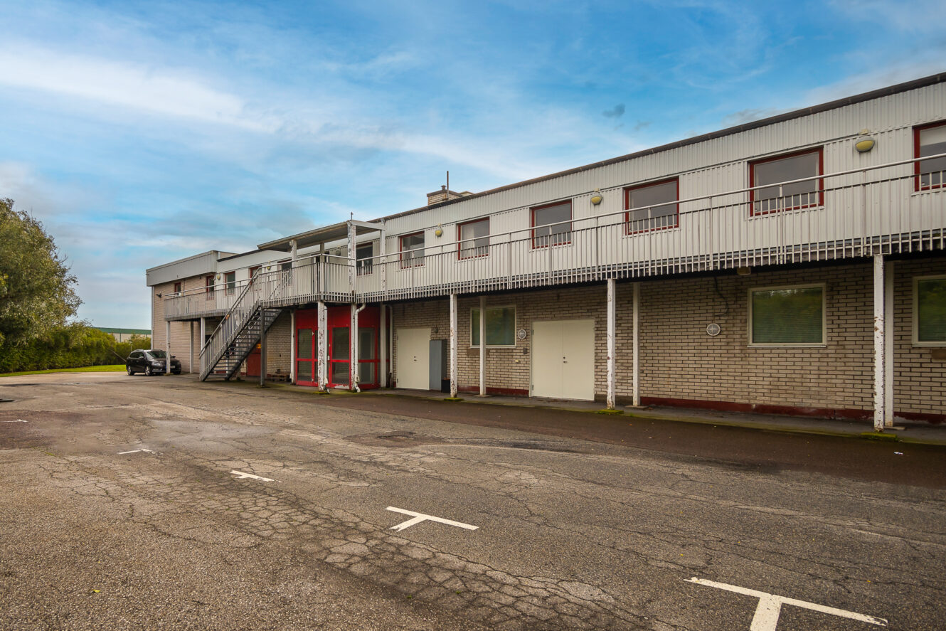Tvåvånings affärsbyggnad med utvändig metalltrappa, röda dörrar och en tom parkeringsplats under en blå himmel.