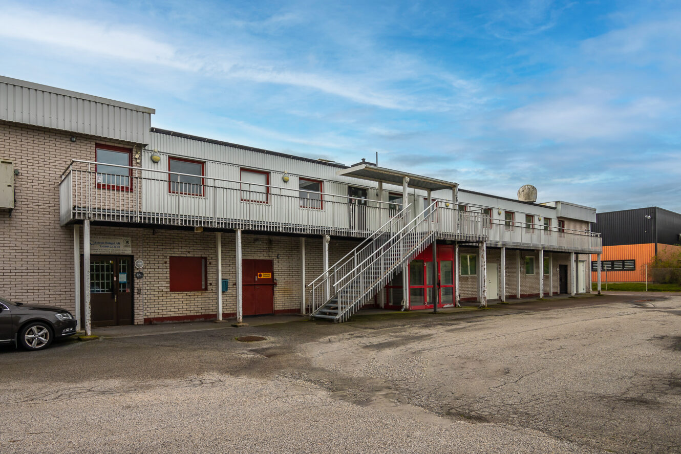 Industribyggnad i två våningar med utvändig metalltrappa, tegelfasad och röda entrédörrar, på en asfalterad tomt under en delvis molnig himmel.