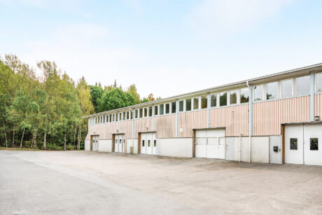 En rad industriella lagerbyggnader med vita garageportar och stora fönster, som ligger intill ett skogsområde under en klar himmel.