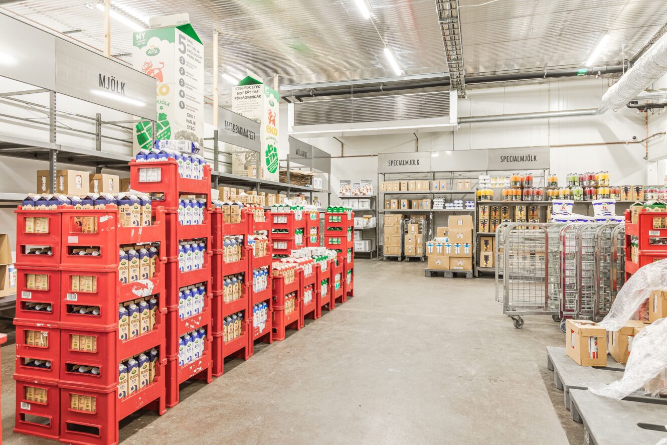 Ett lager med hyllor och röda ställningar fyllda med mjölkkartonger, drycker på flaska och diverse produkter i kartonger på ett betonggolv.