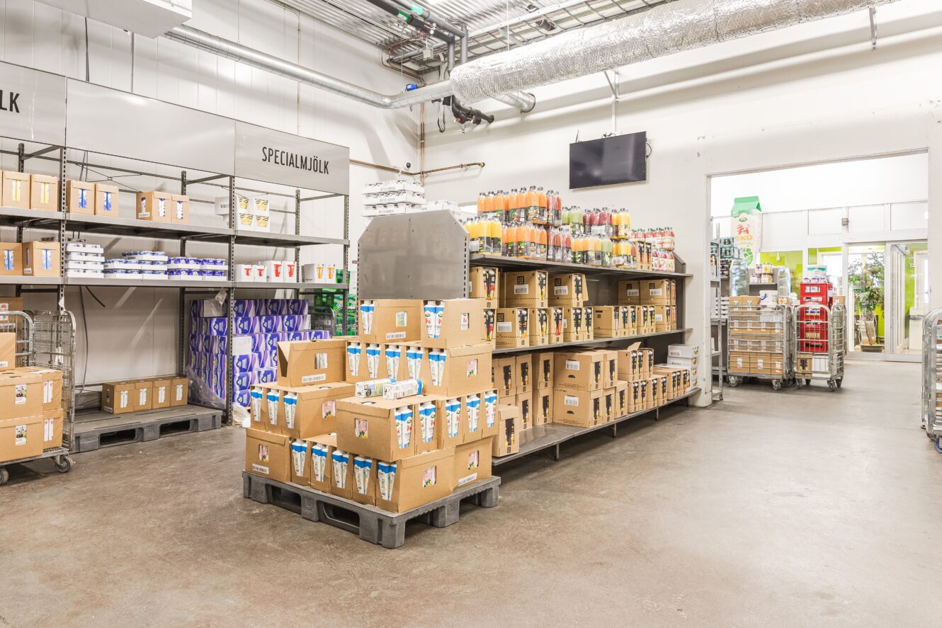 På hyllor och pallar i ett livsmedelsbutikslager står mjölkförpackningar i kartonger och juiceflaskor. Skyltar anger avdelningarna som ”SPECIAL MILK”. Utrymmet är starkt upplyst och man kan se ventilationsöppningar ovanför.