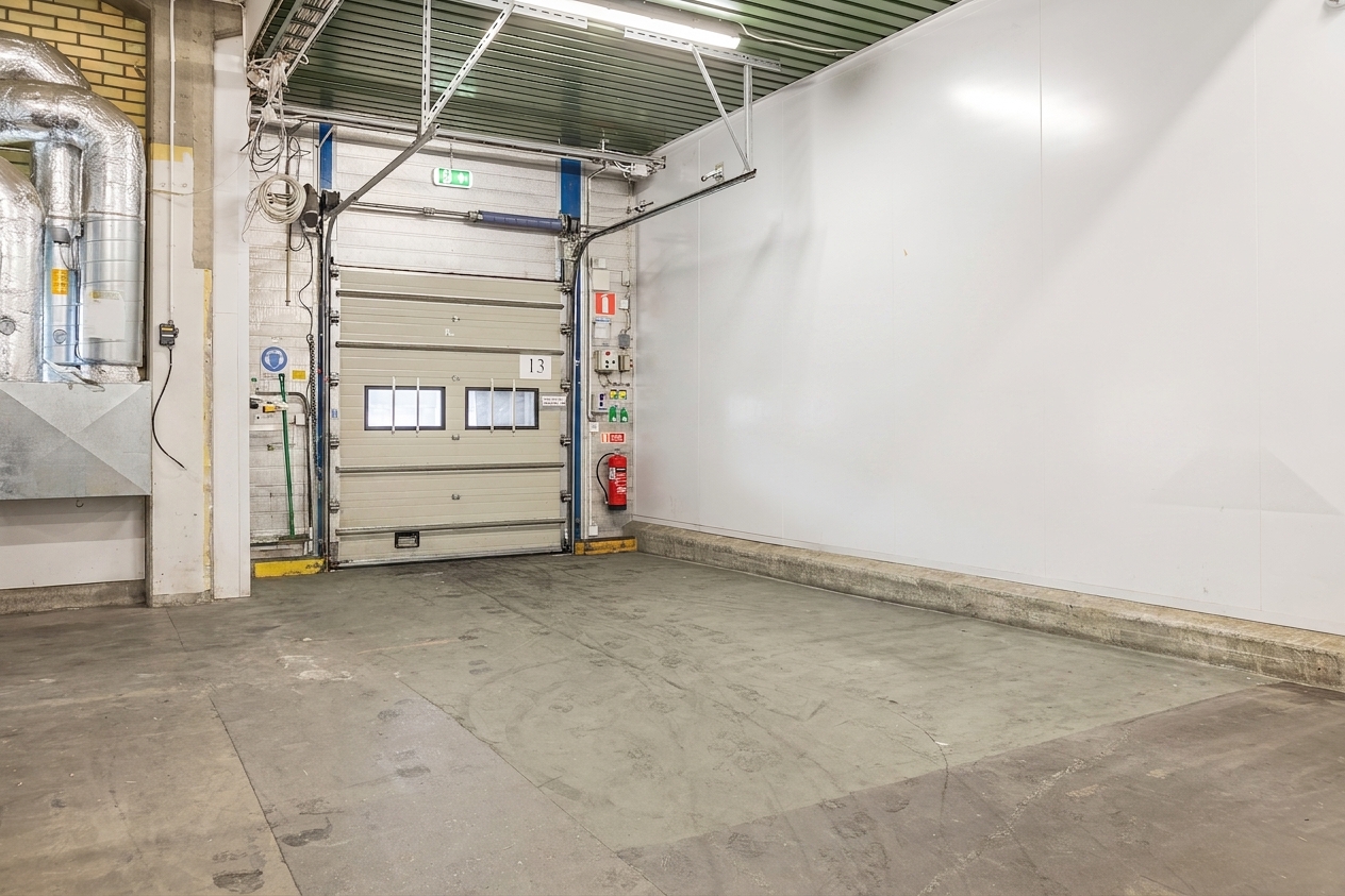 En tom industriell lastkaj med en stängd garageport, en brandsläckare, väggmonterad utrustning och synliga rör på vänster sida.