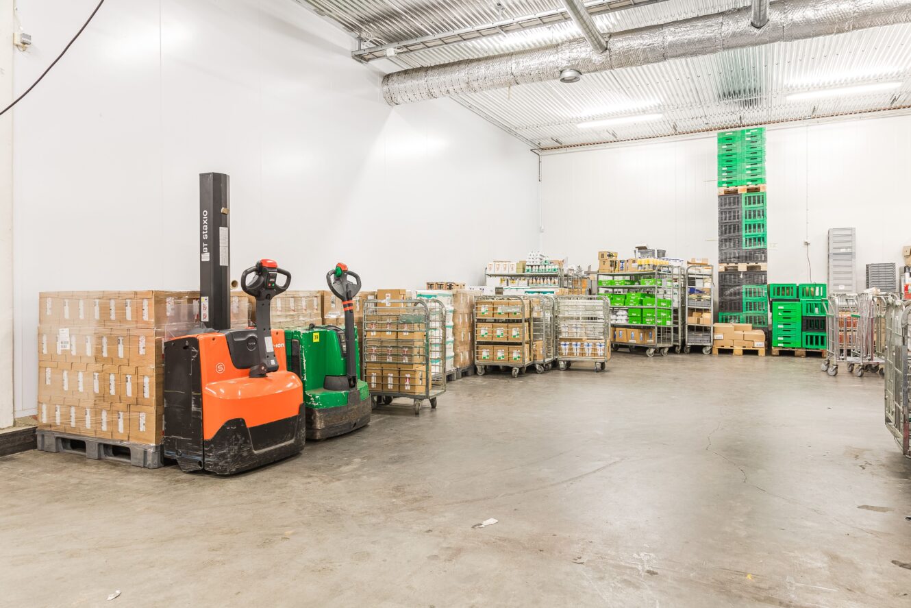 En lagerlokal med staplade pallar, gröna och orangea gaffeltruckar, metallvagnar och plastlådor uppställda mot vita väggar på ett betonggolv.