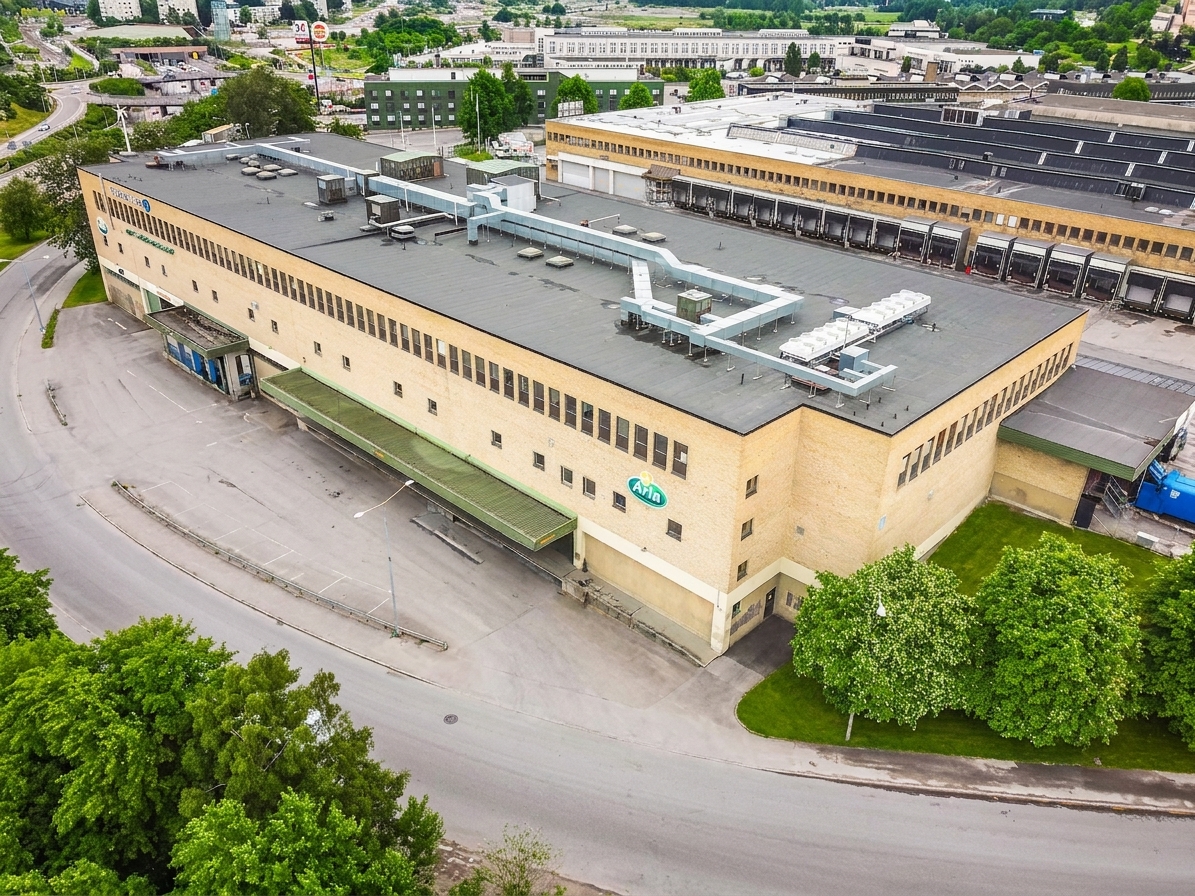Stort beige industriellt lager med platt tak, lastkajer och ventilationsanläggningar, omgivet av asfalterade vägar och grönområden i ett industriområde.