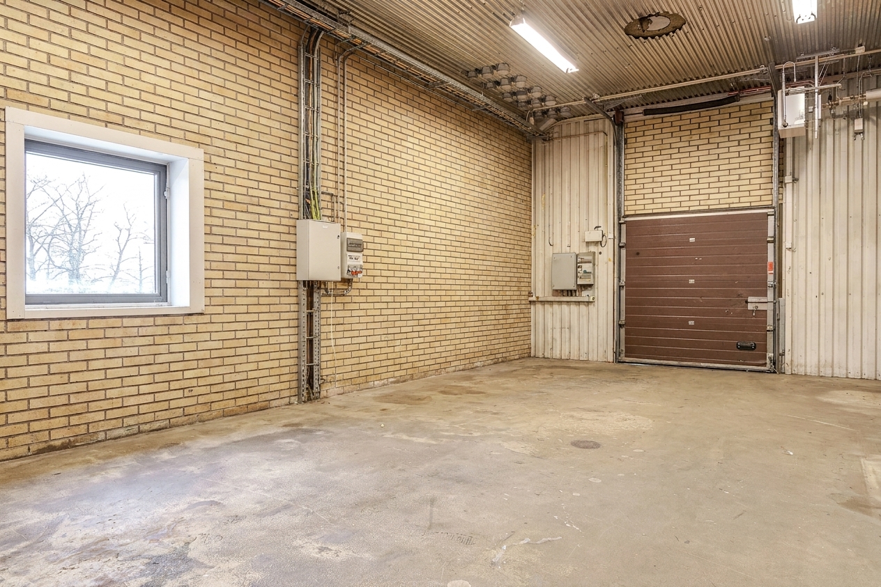 Ett tomt industrigarage med tegelväggar, ett enda fönster, rör som löper längs taket, elskåp och en stängd brun garageport. Betonggolvet har synliga fläckar och märken.