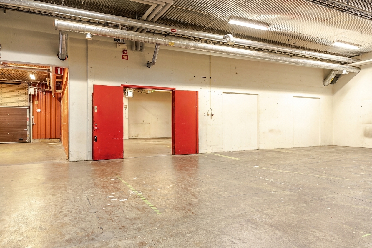 Stort tomt industrilokale med betonggolv, takbelysning, synliga rör, en röd dubbeldörr och en lastdörr i bakgrunden.
