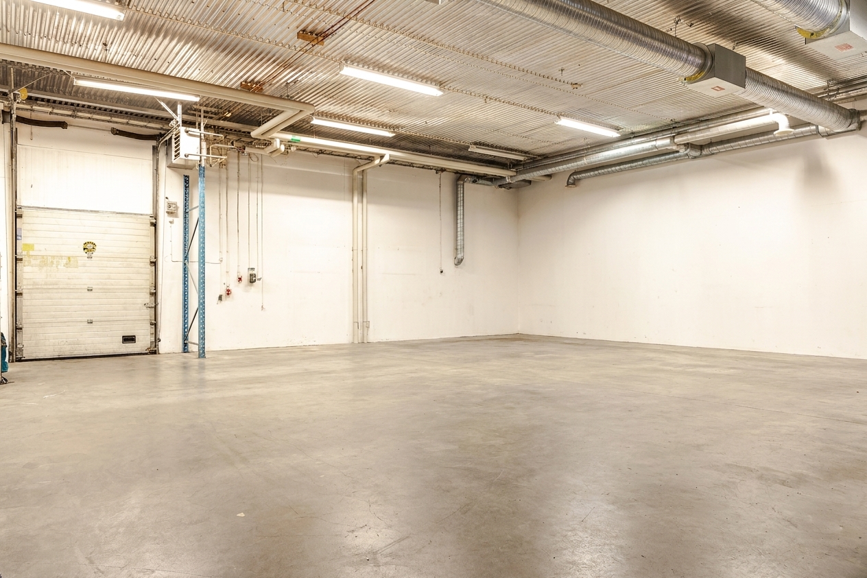 Interiör i ett tomt industrilager med betonggolv, vita väggar, synliga ventilationskanaler och lysrörsbelysning. En stor garageport och rör syns på vänster sida.