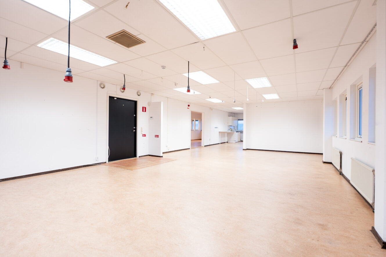 Stort, tomt rum med vita väggar, beige golv, lysrör i taket, hängande kablar och flera fönster och dörrar.