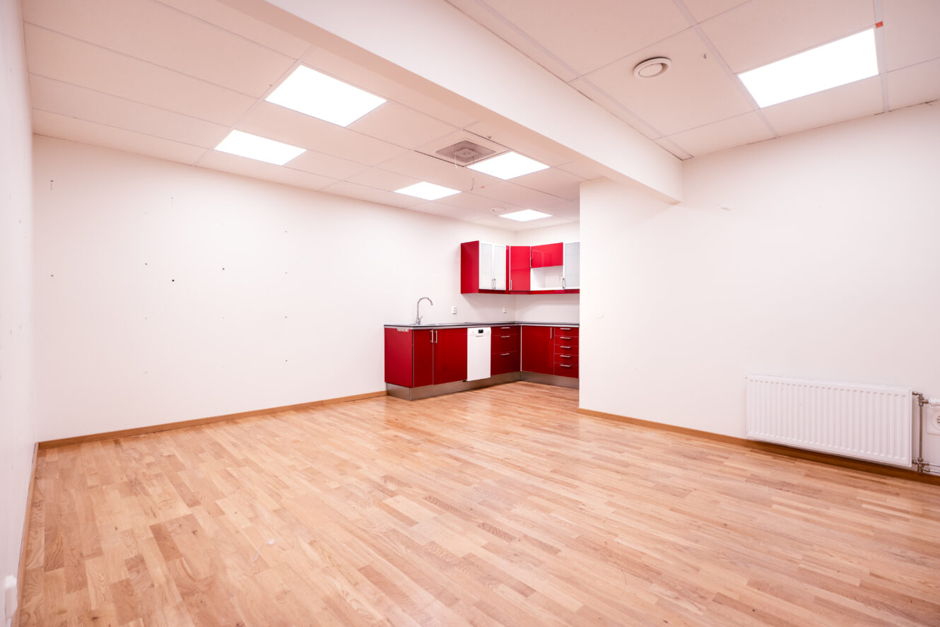 Ett i stort sett tomt rum med ljust trägolv, vita väggar, taklampor och en liten köksdel med röda skåp i hörnet.