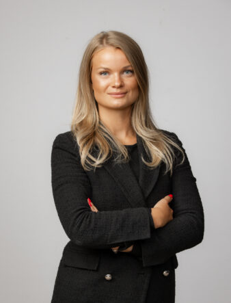 En kvinna med långt blont hår står mot en slät bakgrund, klädd i en svart kavaj med textur och en svart topp, med armarna i kors och ett neutralt ansiktsuttryck.