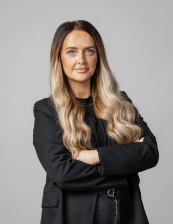 Kvinna med långt vågigt blont hår och svart kavaj står med armarna i kors mot en ljusgrå bakgrund.