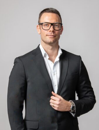 En man i svart kostym, vit skjorta och glasögon står mot en ljus bakgrund, vänd mot kameran och med ena handen i kavajen.