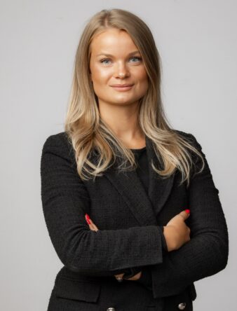 En kvinna med långt blont hår, klädd i en svart kavaj, står med armarna i kors mot en ljus bakgrund.