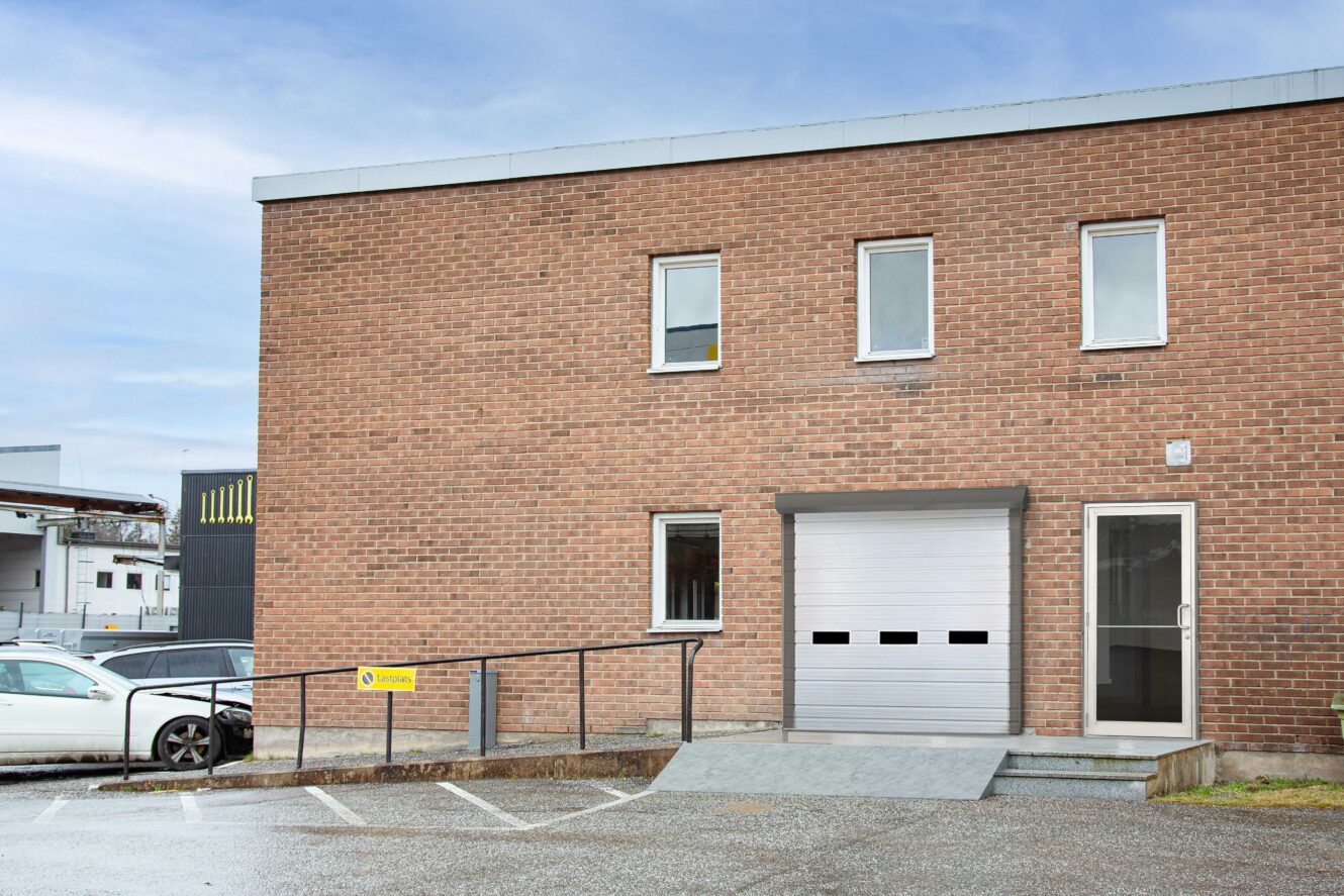En industribyggnad i tegel med tre fönster, en garageport med ramp, en entrédörr i glas och en parkerad vit bil utanför.