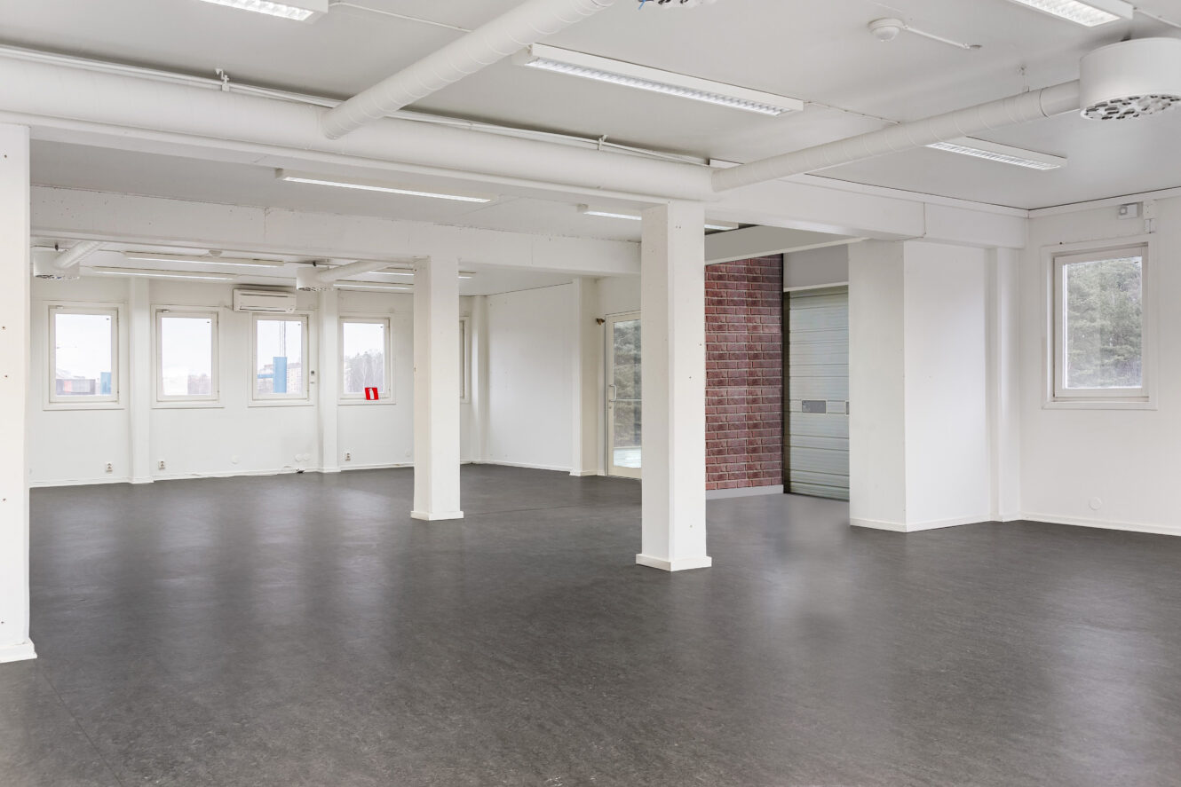 Tomt, modernt rum med öppen planlösning, vita väggar, flera fönster, synliga rör, stödpelare och mörkt golv. En delvis stängd garageport syns i bakgrunden.