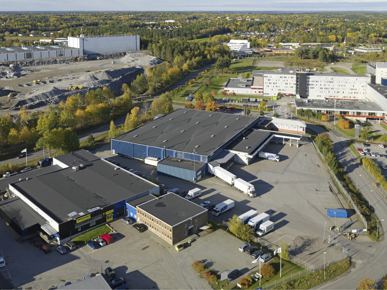 Flygfoto över ett stort industriellt lagerkomplex med lastkajer, parkerade lastbilar, bilar och närliggande kontorsbyggnader omgivna av träd och vägar.