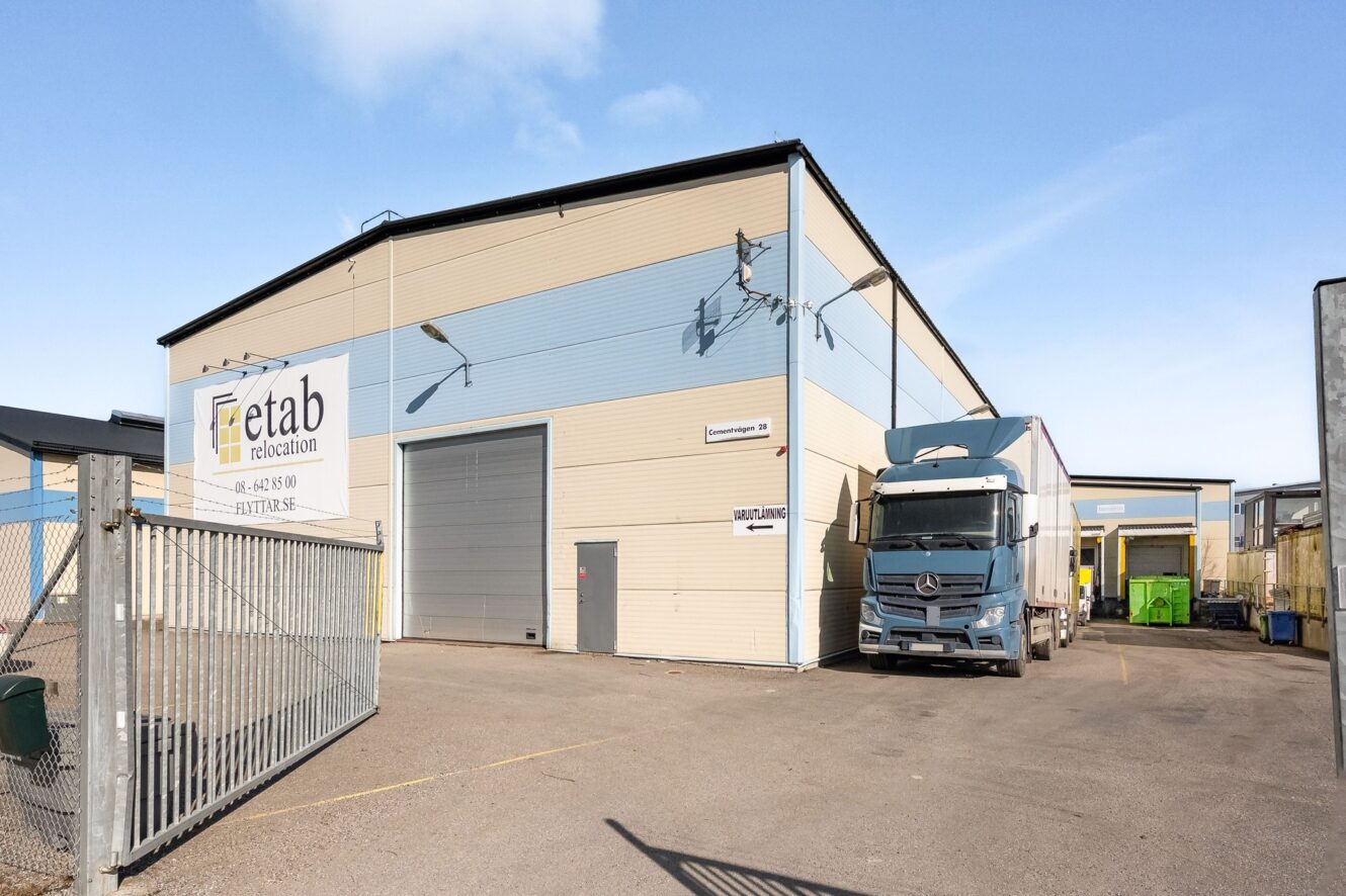 En stor industrihall med en etab relocation-skylt, en stängd garageport, övervakningskameror och en parkerad blåvit lastbil utanför entrén.