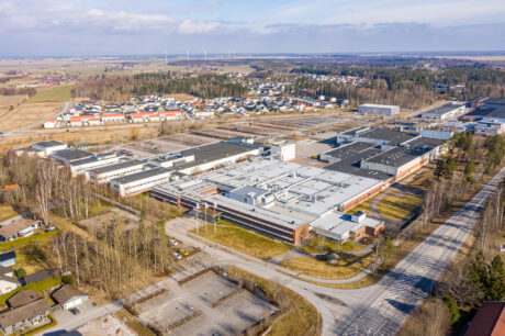 Flygfoto över en stor industrianläggning omgiven av bostadsområden, träd och öppna fält under en delvis molnig himmel.