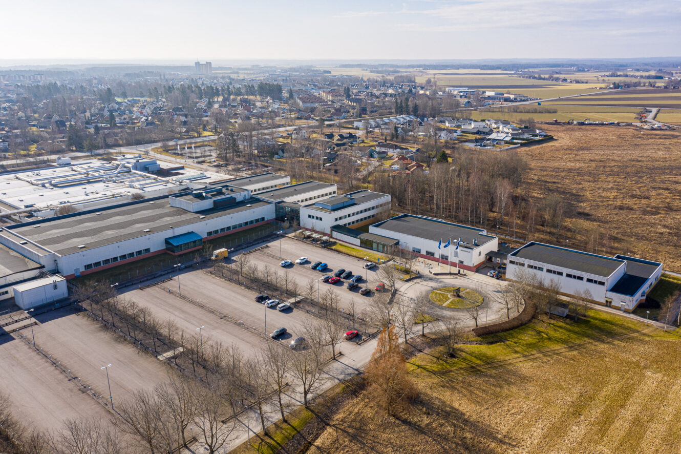 Flygfoto över ett stort industri- eller skolkomplex med parkeringsplats, omgivet av fält och ett förortsbostadsområde i bakgrunden.