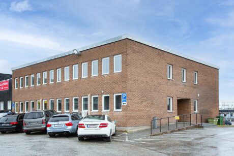 En tvåvånings kontorsbyggnad i tegel med flera fönster, en ramp och parkerade bilar framför under en molnig himmel.
