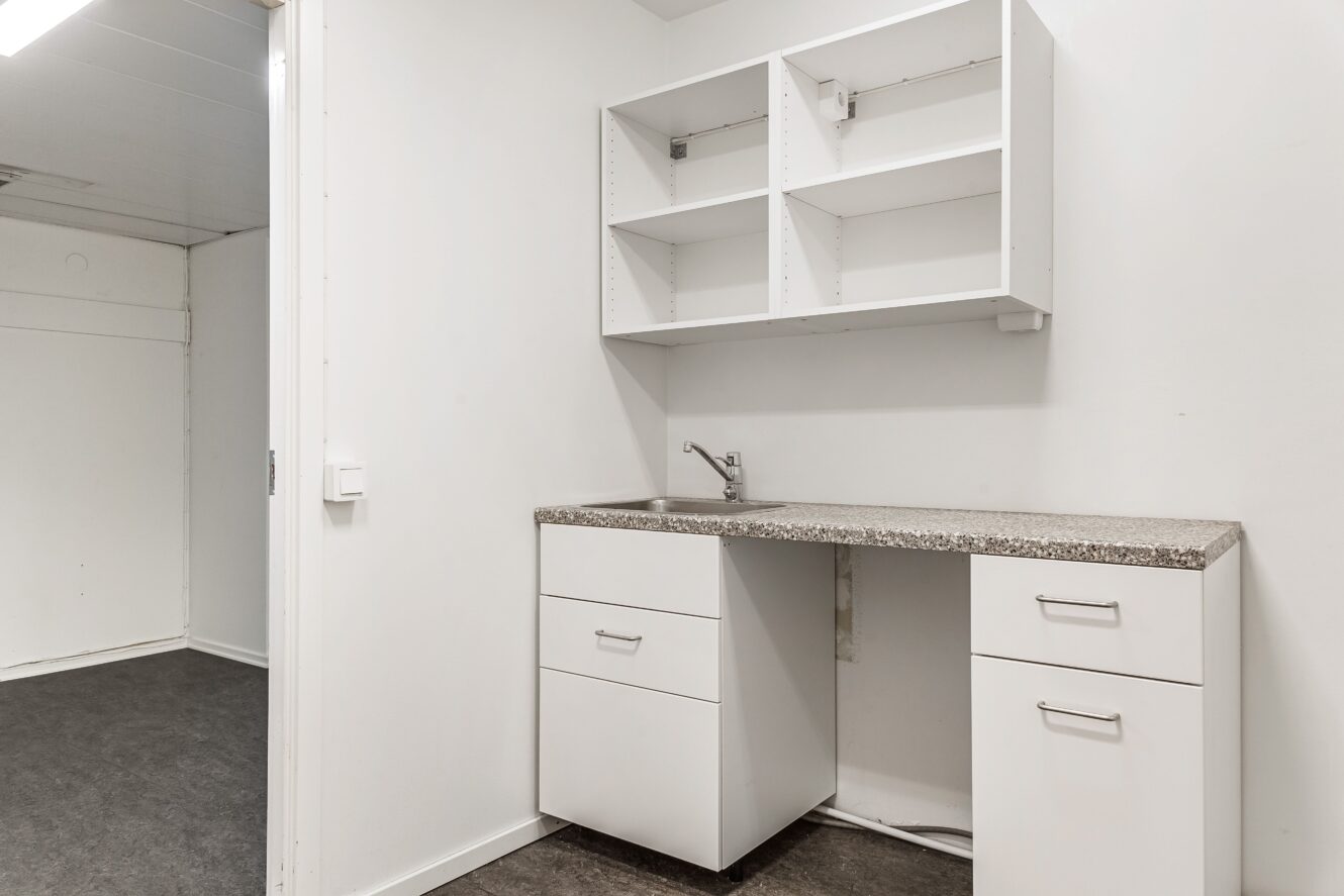 Ett litet, minimalistiskt pentry med vita skåp, öppna hyllor, en enda kran och en bänkskiva i granit i ett vanligt vitt rum.