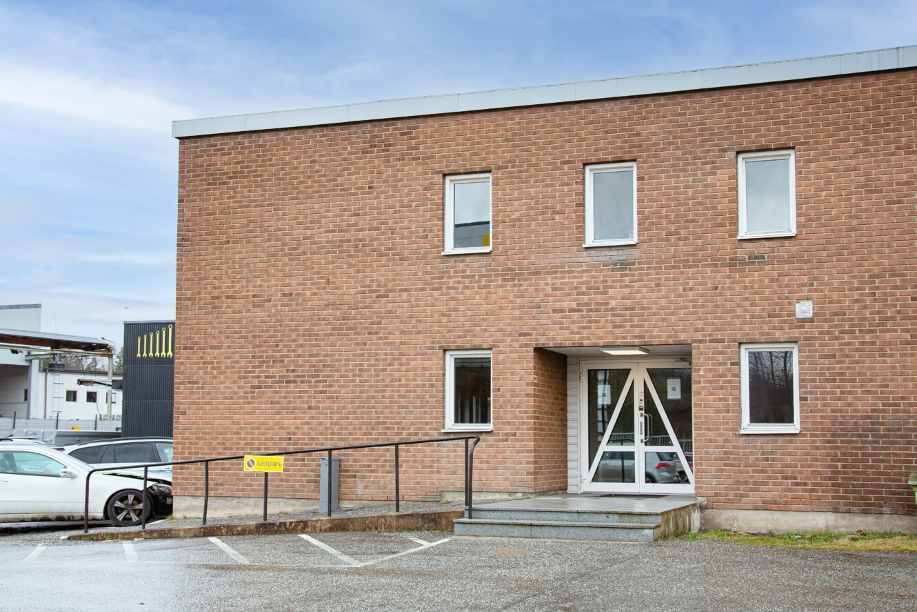 En tvåvånings tegelbyggnad med flera fönster och en huvudingång med ramp och trappor utanför. En vit bil står parkerad i närheten.