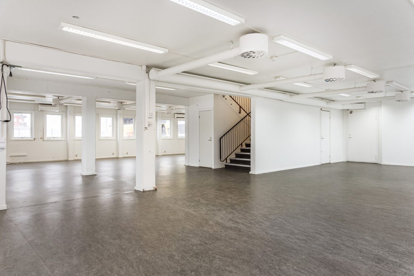 Rymligt, tomt rum med vita väggar, stora fönster, lysrör i taket och en trappa som leder upp till en övre våning.