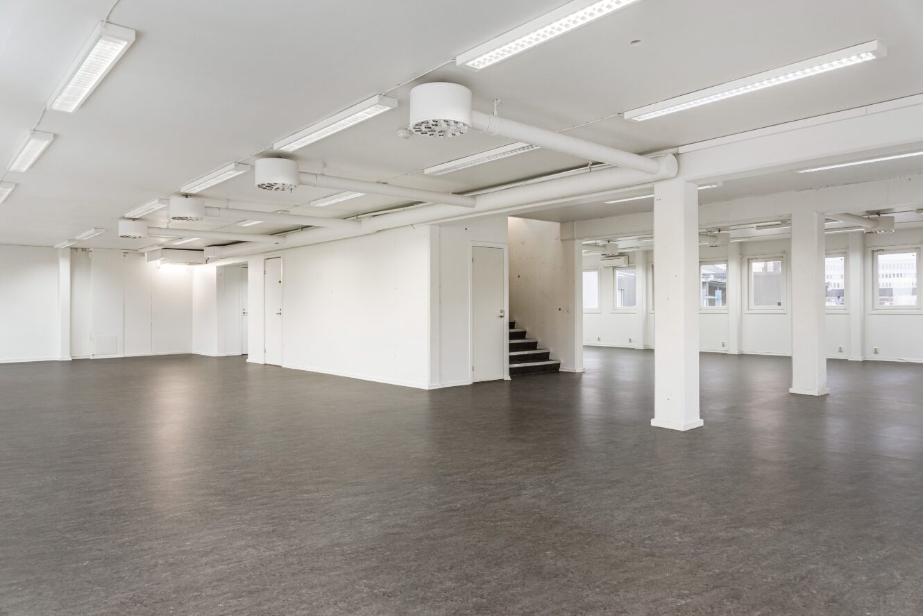 Stort tomt kontorsutrymme med vita väggar, grått golv, lysrör i taket, synliga rör och en liten trappa som leder till ett övre plan.