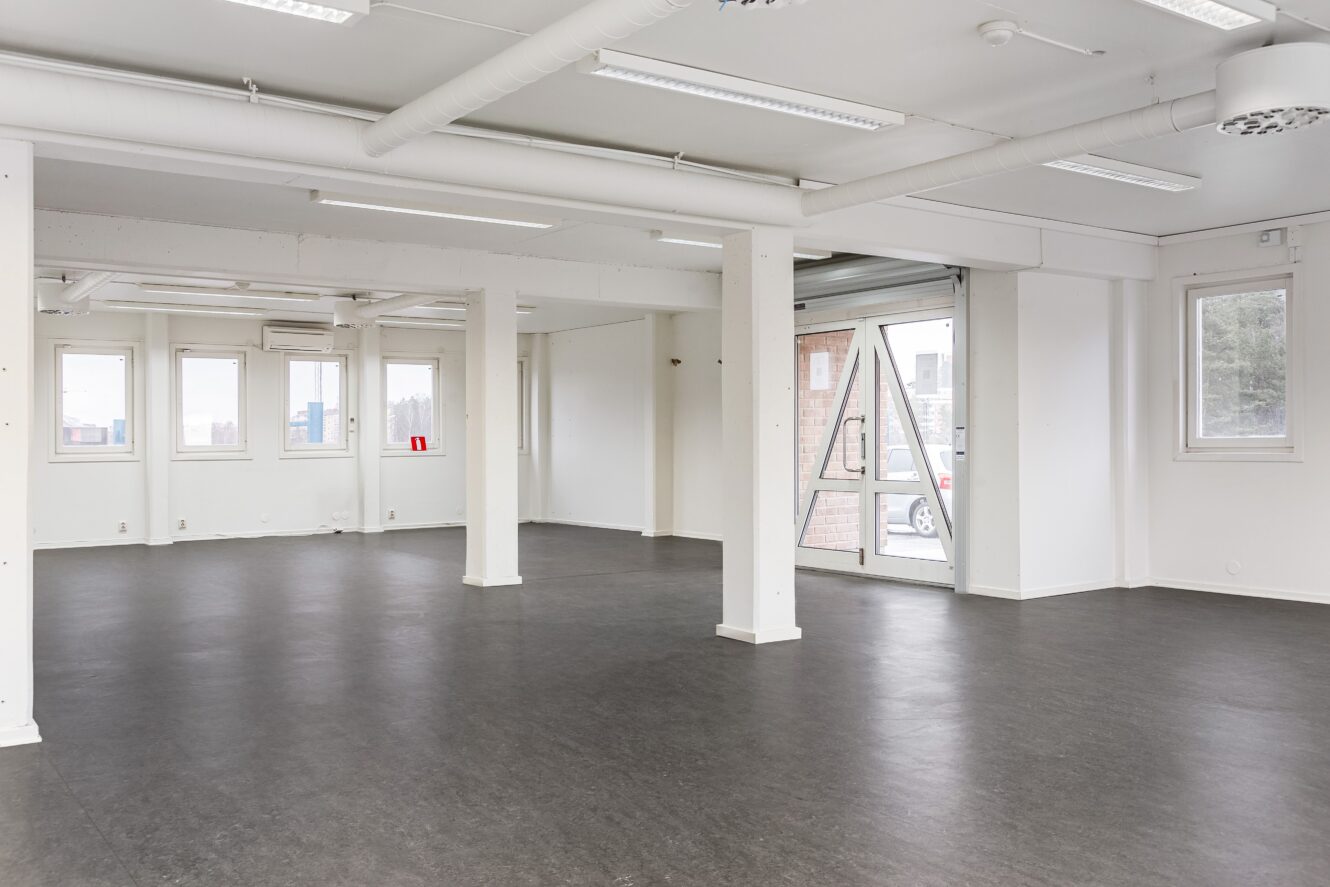 Tomt, omöblerat rum med vita väggar, stora fönster och mörkt golv; synliga rör och pelare syns i hela utrymmet.