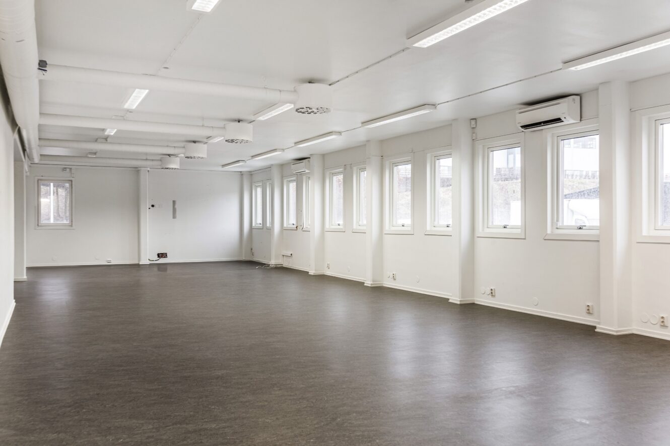 Stort, tomt kontorsutrymme med vita väggar, flera fönster, mörkt golv, taklampor och synliga kanalsystem.