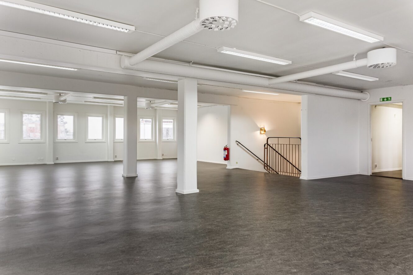 Rymligt, tomt kontorsrum med vita väggar, stora fönster, mörkt golv, taklampor och en trappa med svart räcke.