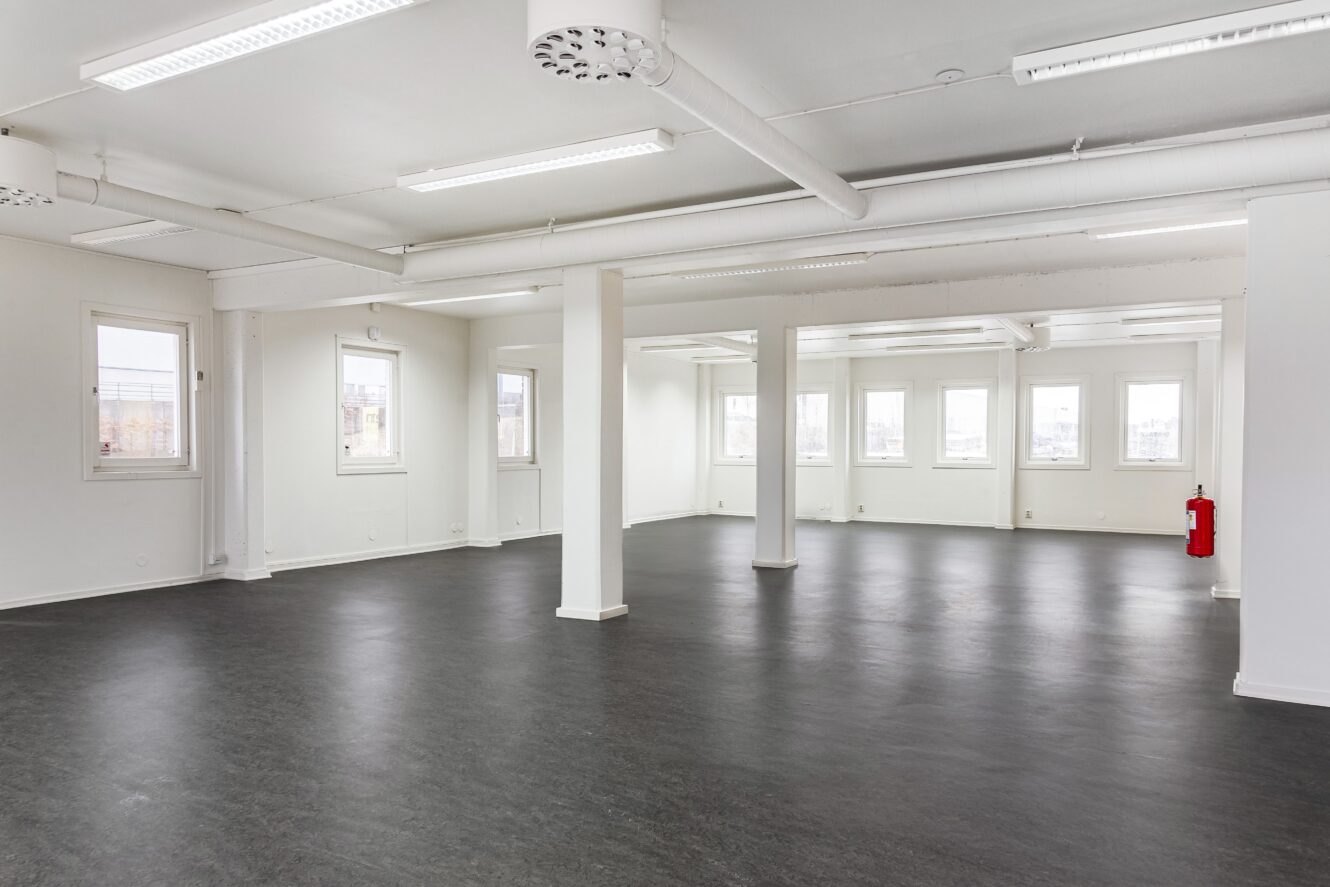 Stort tomt kontorsrum med vita väggar, flera fönster, svart golv, rör i taket och en röd brandsläckare i hörnet.