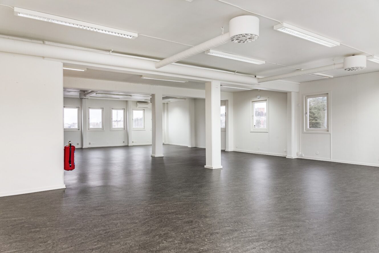 Tomt, öppet kontorslandskap med vita väggar, stora fönster, grått golv, taklampor och en röd brandsläckare monterad på en pelare.