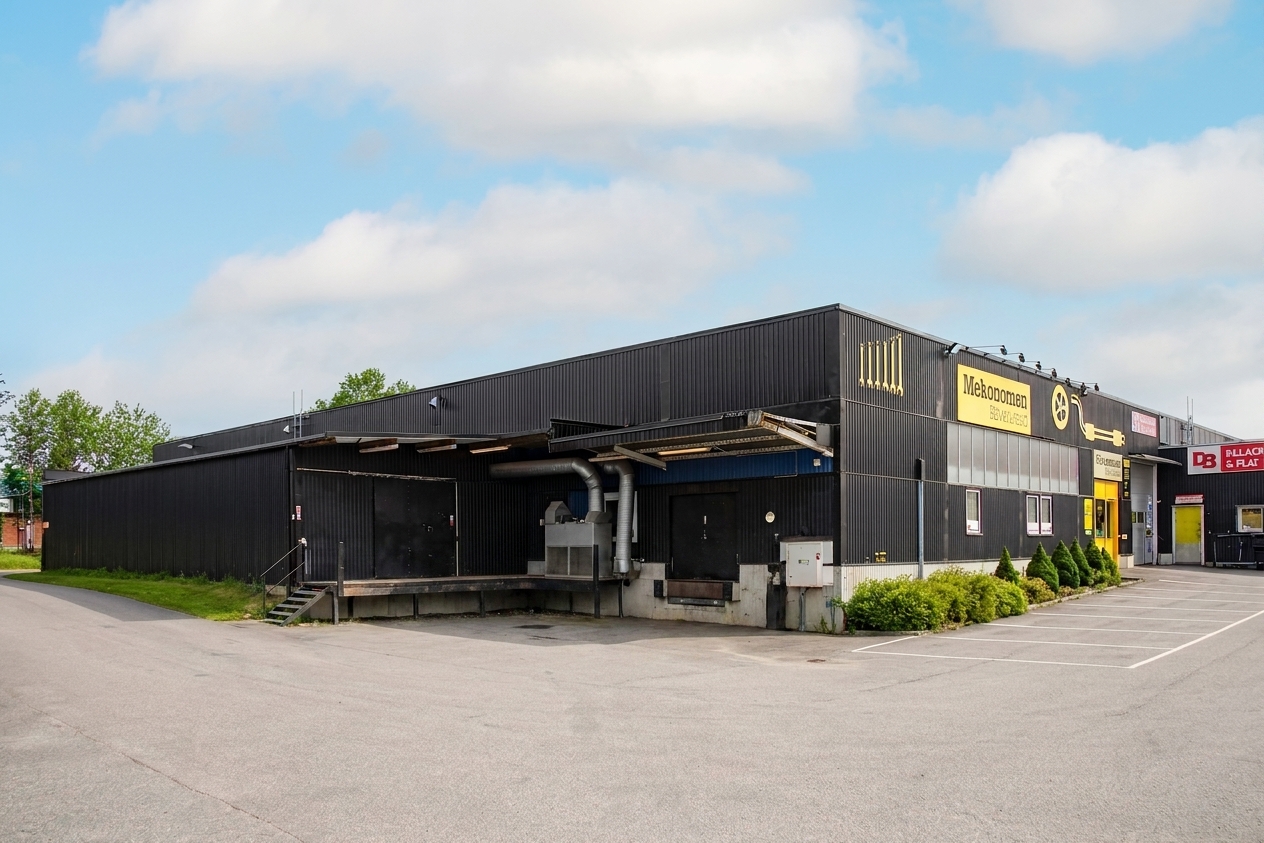 En stor svart industribyggnad med gula skyltar, omgiven av en asfalterad tomt och lite grönska, under en delvis molnig himmel.