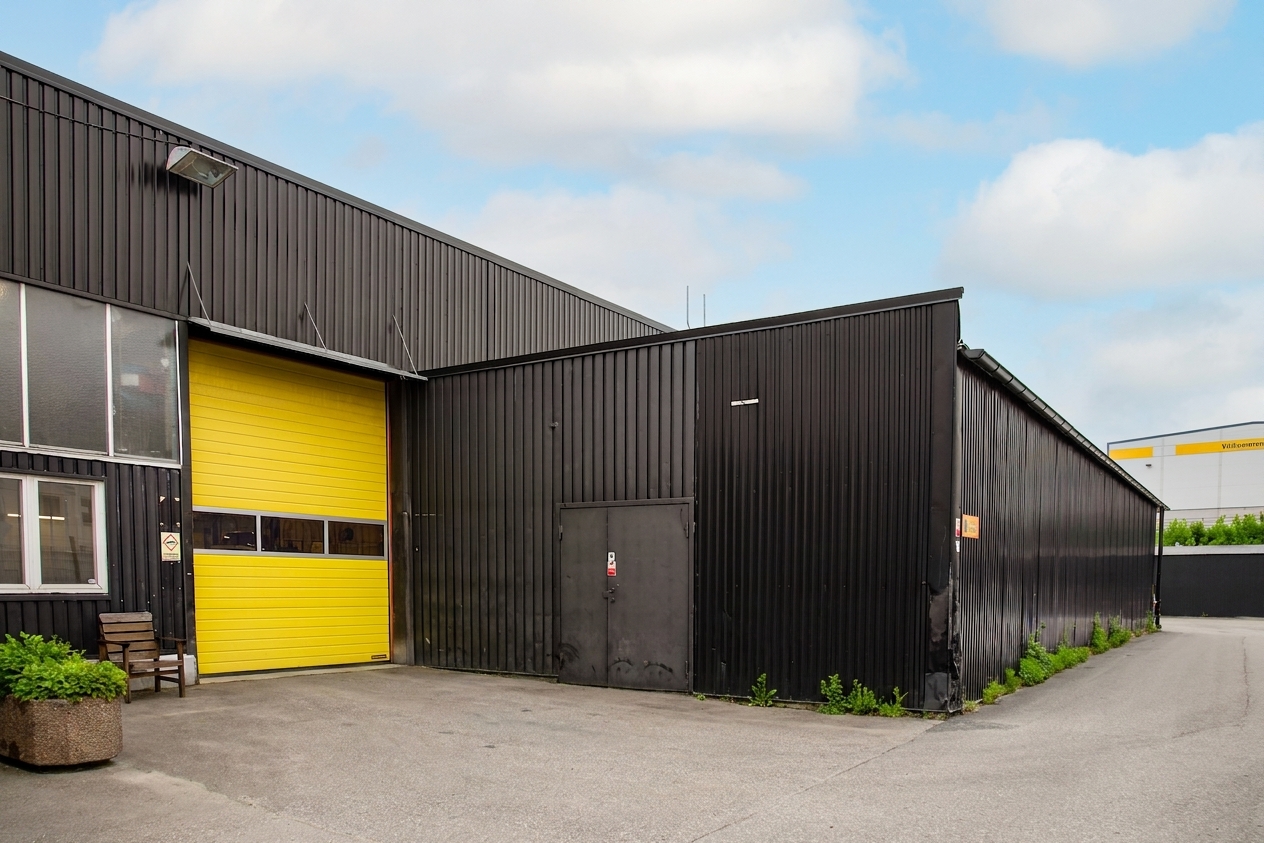 Ett svart industrilager med en stor gul garageport och en sid ingång, beläget på en asfalterad tomt under en delvis molnig himmel.