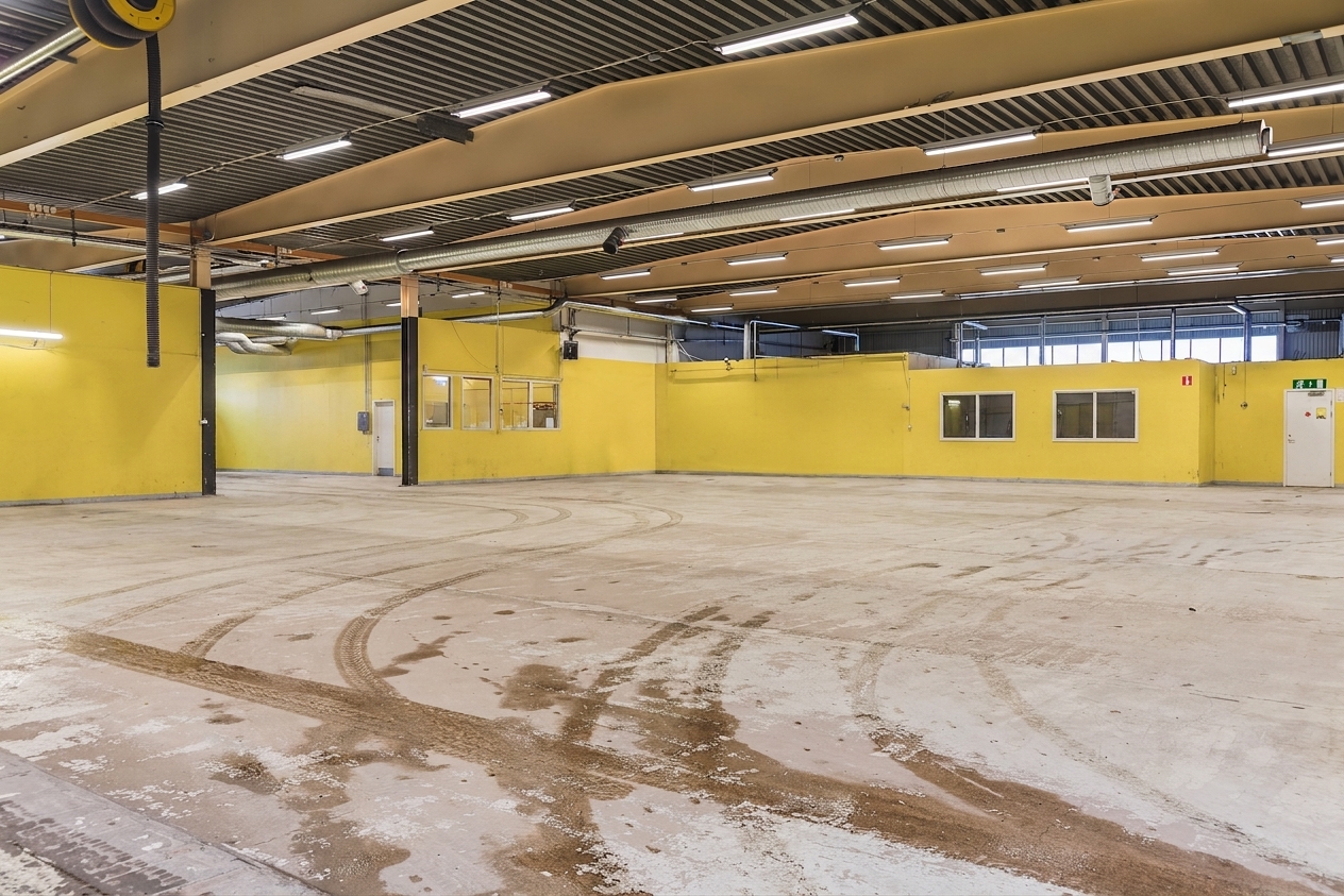 Stort tomt lagerutrymme med gula väggar, högt i tak, synliga ventilationskanaler och däckspår på betonggolvet. Flera fönster och dörrar syns längs väggarna.