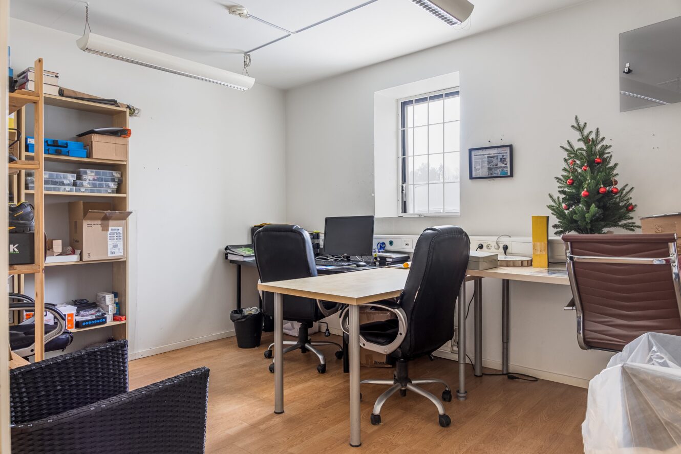 Ett litet kontor med två skrivbord och stolar, datorskärmar, hyllor med lådor, en papperskorg och en pyntad minijulgran som står bredvid fönstret.